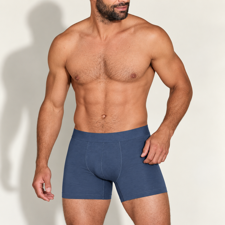 Gespierde man met baard draagt een blauwe boxershort met langere pijp, voorkant, op een lichte achtergrond met schaduw. 1