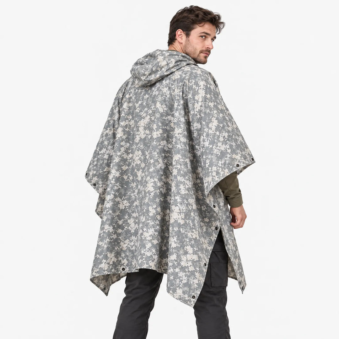 Achterkant van de poncho in grijs-beige pixelcamouflage, gedragen door een model dat over zijn schouder kijkt, met capuchon en drukknopen. 4