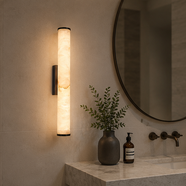 Wandlamp met gemarmerde lichtbuis en zwarte afwerking naast een ronde spiegel boven een marmeren wastafel met zeepdispenser en vaas. 4