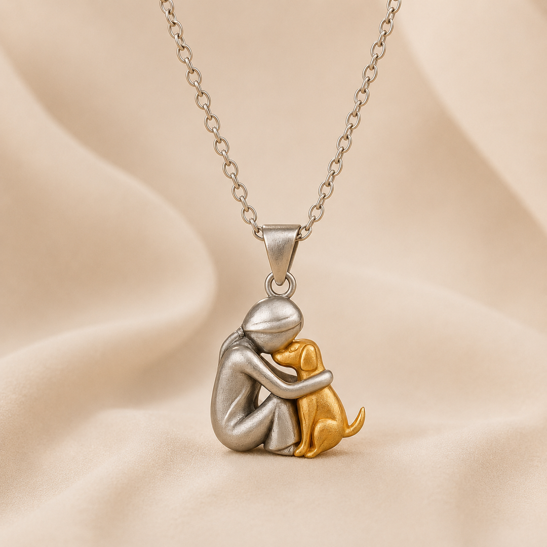 Hanger van een zittende figuur in mat zilverkleur die een goudkleurige hond omarmt, hangend aan een schakelketting op beige stof. 1