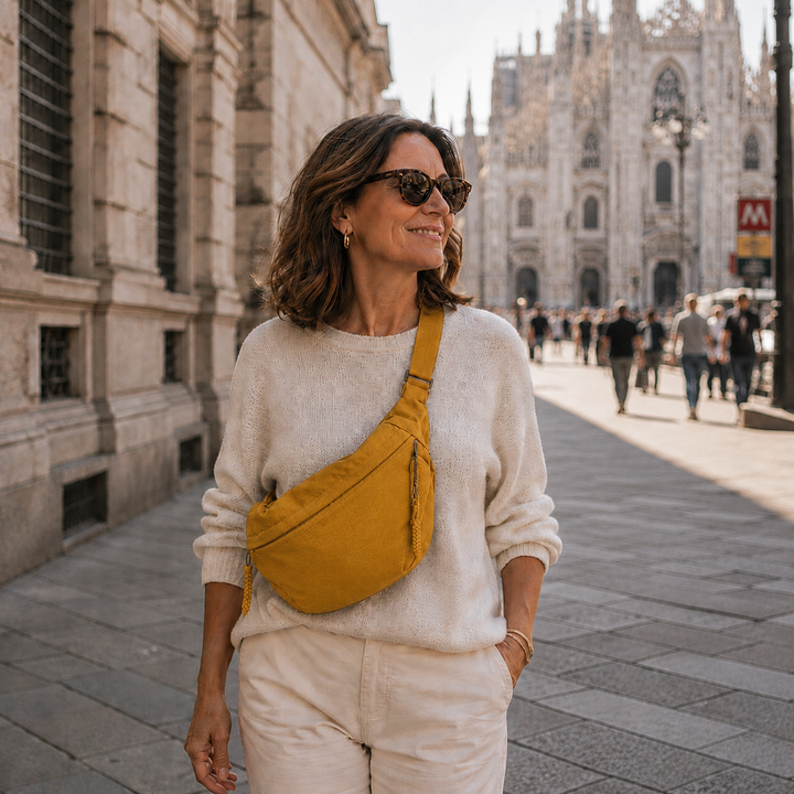 Vrouw met zonnebril en crèmekleurige trui draagt de okergele tas als crossbody op een zonnig plein in een oude stad. 4