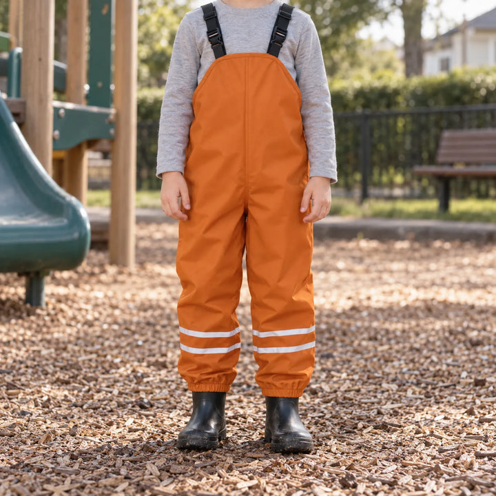 Kind in oranje tuinbroek met verstelbare bretels staat op een speelplaats met houtsnippers, met grijs shirt en zwarte rubberlaarzen. 3