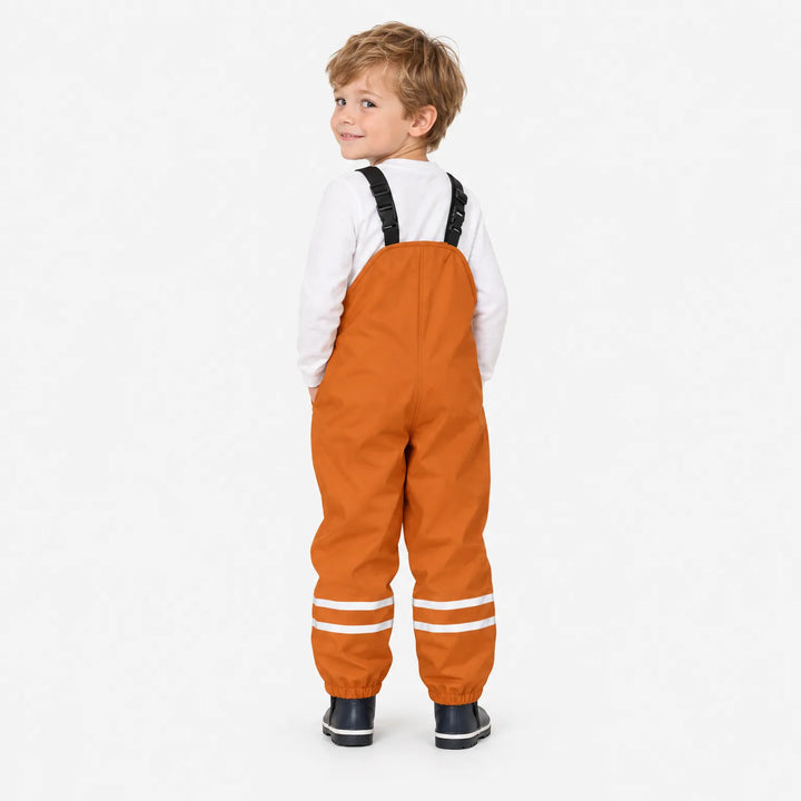 Achterkant van oranje tuinbroek gedragen door een lachend jongetje dat over zijn schouder kijkt, met wit shirt en donkere laarzen. 4