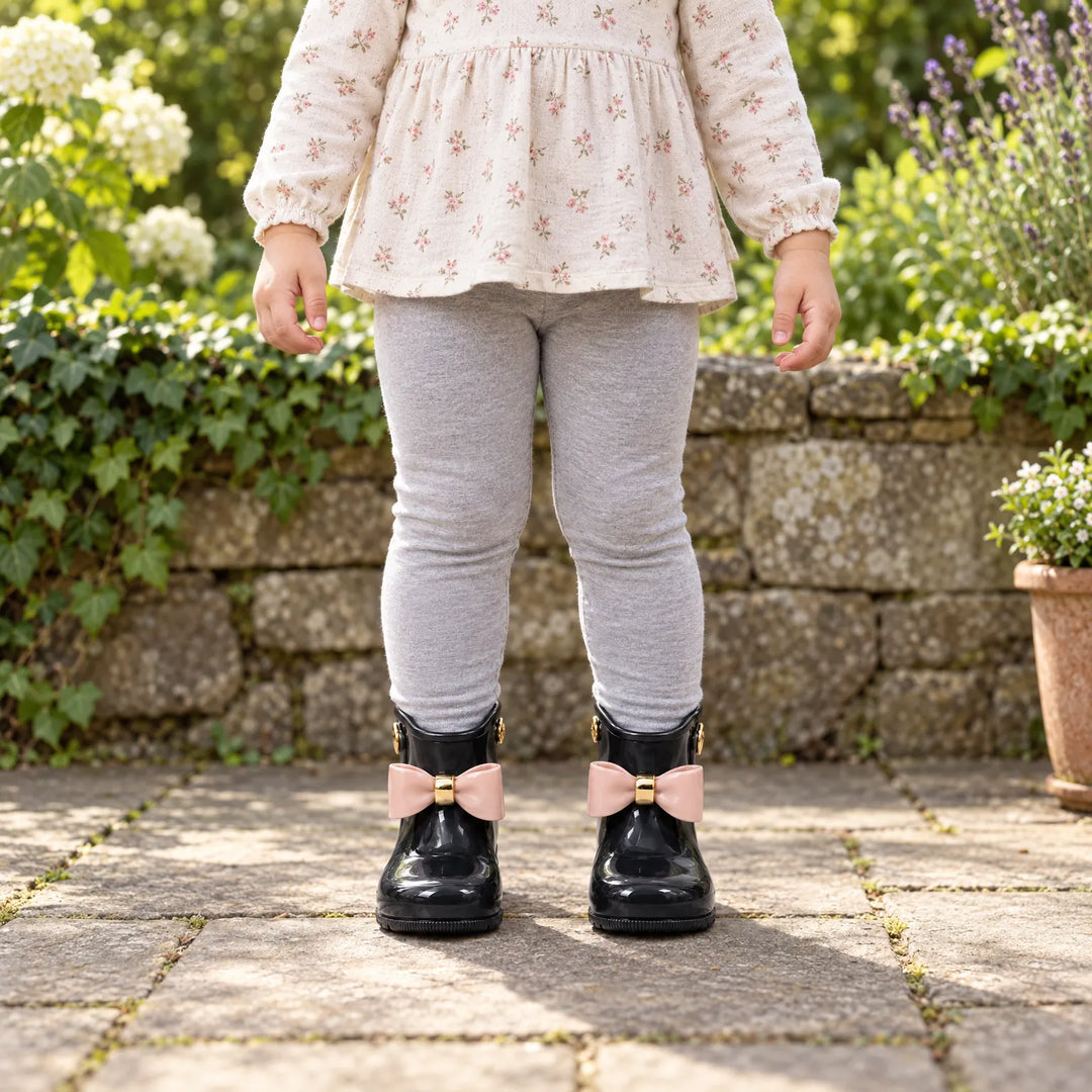 Peuter in bloemenblouse en grijze legging draagt zwarte glanzende laarsjes met roze strik in een tuin. 3