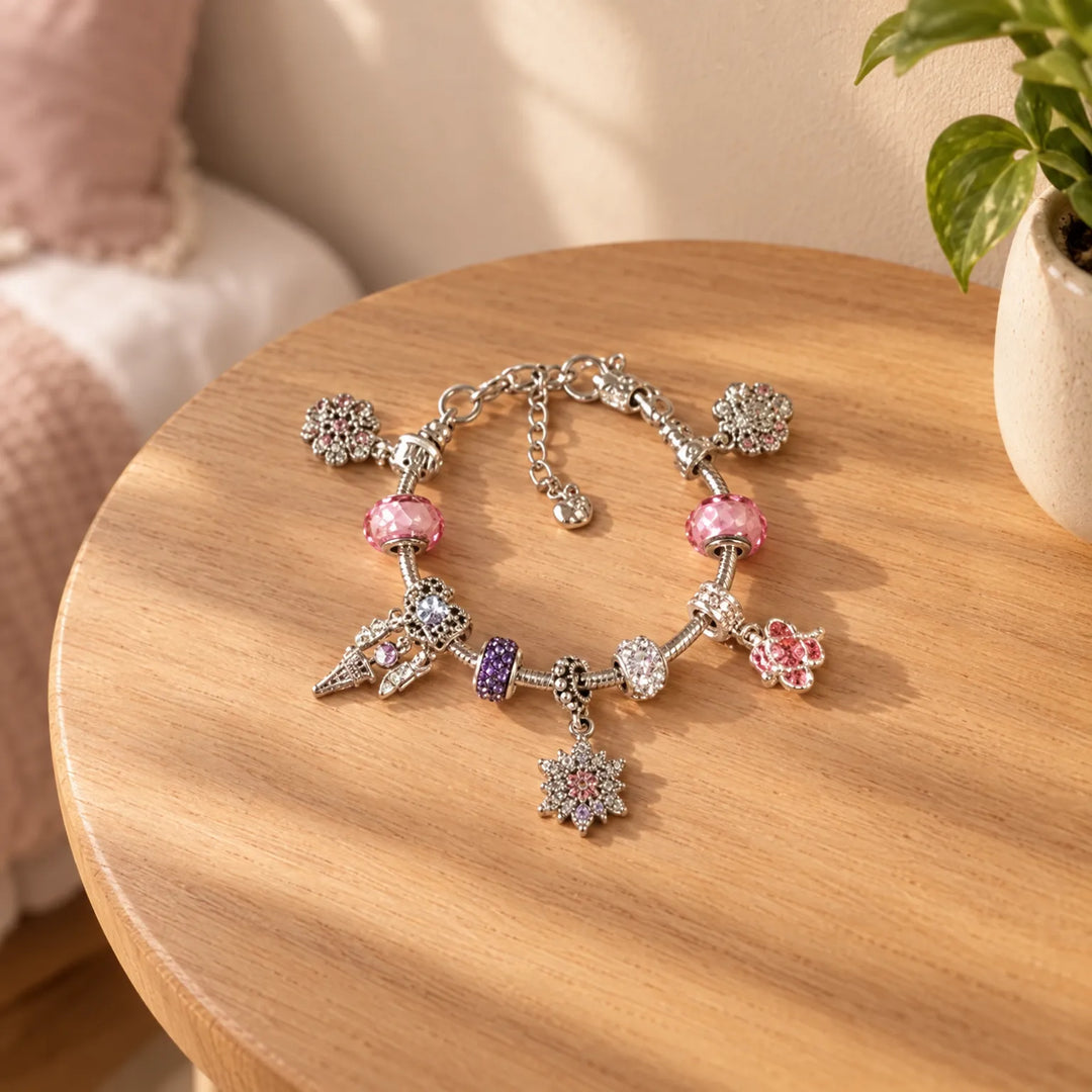 Afgewerkt zilverkleurig bedelarmband met roze en paarse kralen en sneeuwvlok- en bloembedels, liggend op een rond houten tafeltje naast een plant. 1
