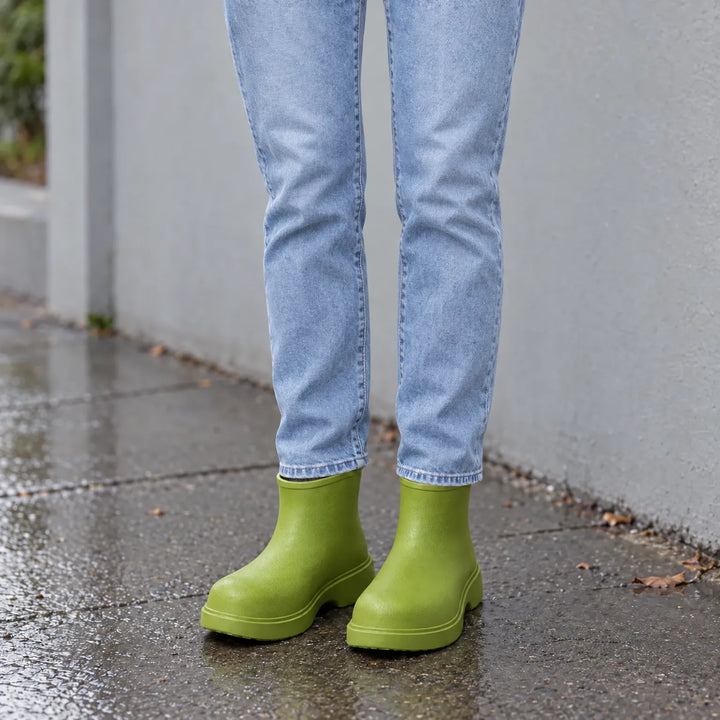 Groene enkellaarzen gedragen met lichtblauwe jeans op een natte stoep, zicht op alleen de onderbenen. 1
