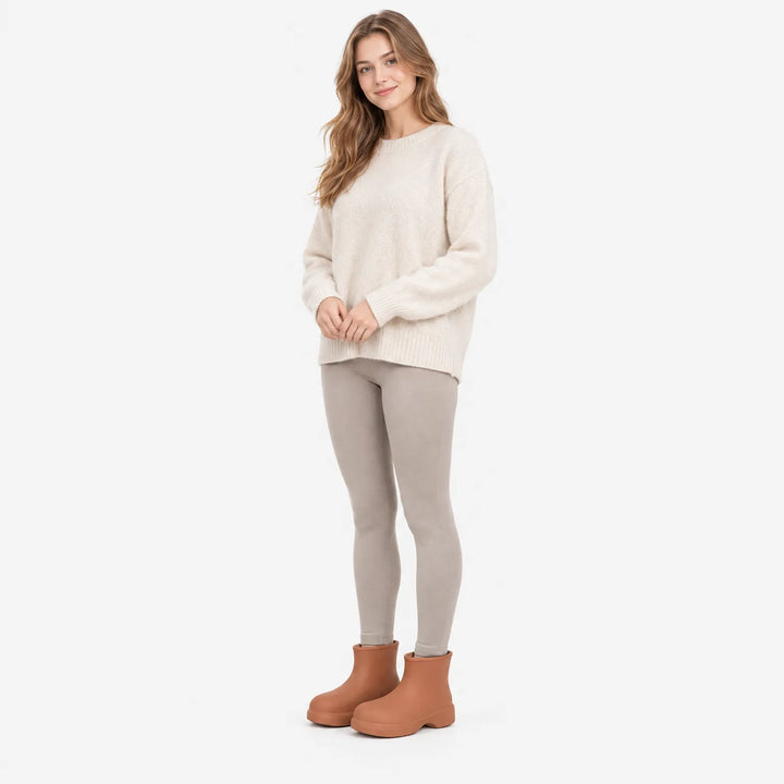 Lachende vrouw in crèmekleurige trui en beige legging, gekleed met bruine enkellaarzen, volledig model op witte achtergrond. 4