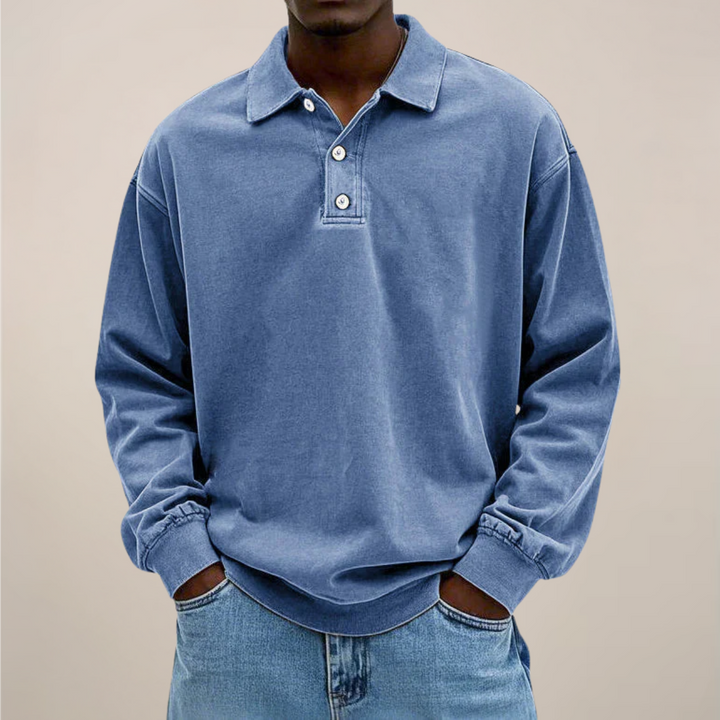Model draagt het poloshirt in blauw, voorkant met drie knopen, handen in de zakken van een spijkerbroek, losse pasvorm. 1