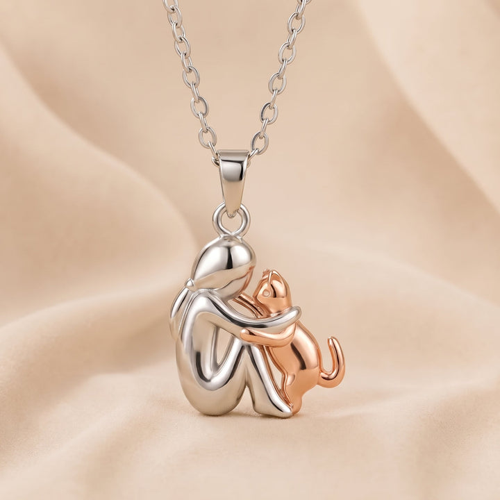 Hanger van een zilverkleurige figuur met een roségoudkleurige kat die haar knuffelt, aan een zilverkleurige ketting op beige stof. 4