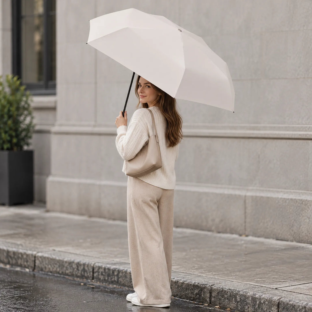 Vrouw in beige kleding kijkt over haar schouder en houdt een geopende crèmewitte paraplu vast, op een natte stoep voor een grijze gevel. 1
