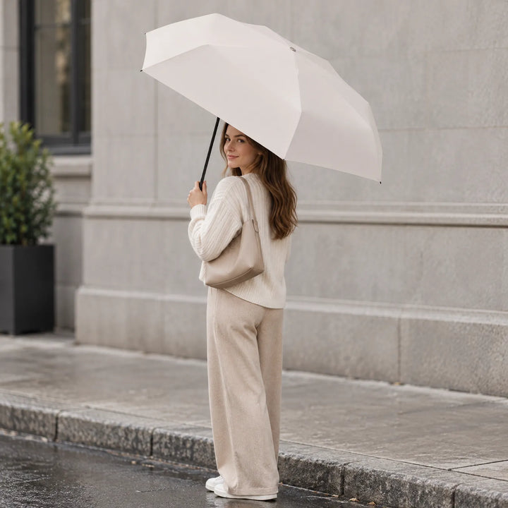 Vrouw in beige kleding kijkt over haar schouder en houdt een geopende crèmewitte paraplu vast, op een natte stoep voor een grijze gevel. 1