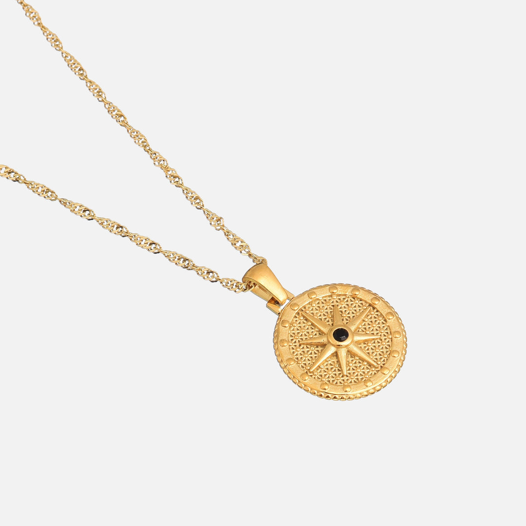 Ronde gouden hanger met kompasster en zwart steentje, schuin van bovenaf gefotografeerd, met de gedraaide ketting op een lichte achtergrond. 2