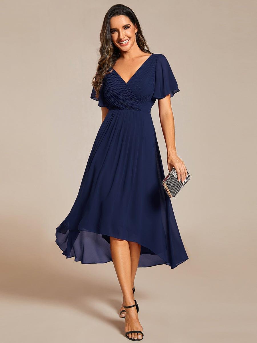 Model in marineblauwe chiffon jurk met gedrapeerde V-hals en wijde mouwtjes, vooraanzicht, met glinsterend clutchtasje in de hand. 1