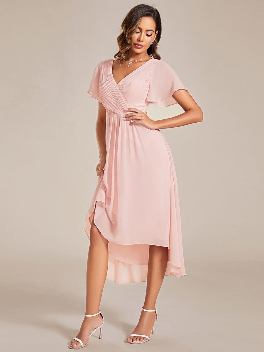 Roze jurk schuin van voren, model tilt licht de rok op zodat de asymmetrische, gelaagde chiffon zoom zichtbaar wordt. 19