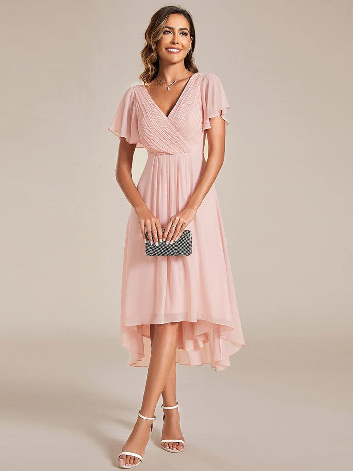 Model in roze chiffon jurk met V-hals en fladdermouwen houdt een zwart clutchtasje voor zich, vooraanzicht. 15