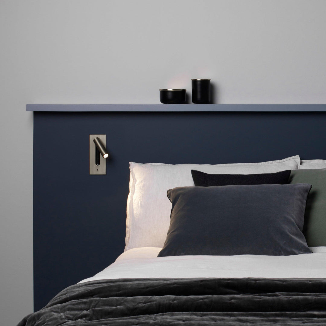 Wandlamp met brons afwerking boven een bed met donkerblauw hoofdbord en grijze kussens, spot aan 30