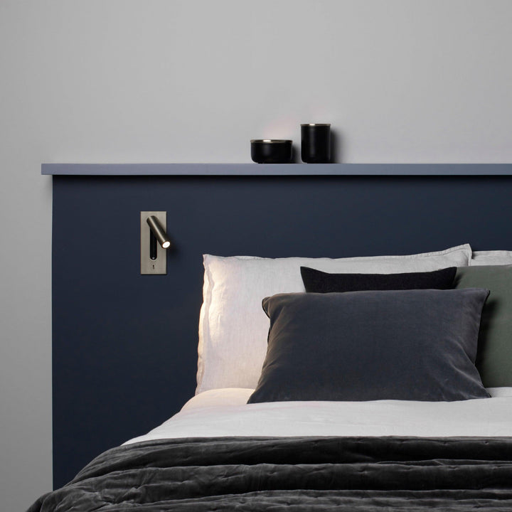 Wandlamp met brons afwerking boven een bed met donkerblauw hoofdbord en grijze kussens, spot aan 30