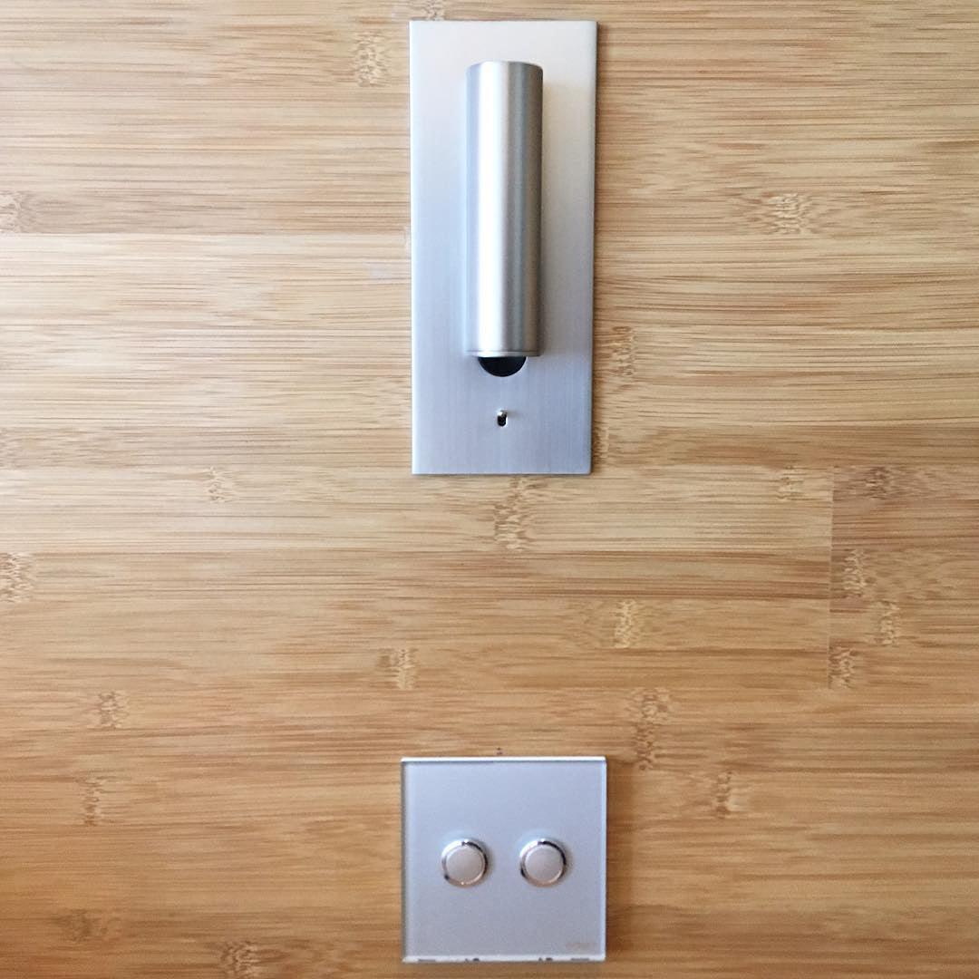 Aluminiumkleurige lamp met rechtopstaande cilinder, uitgeschakeld, op bamboehouten wand boven een dubbele schakelaar 34