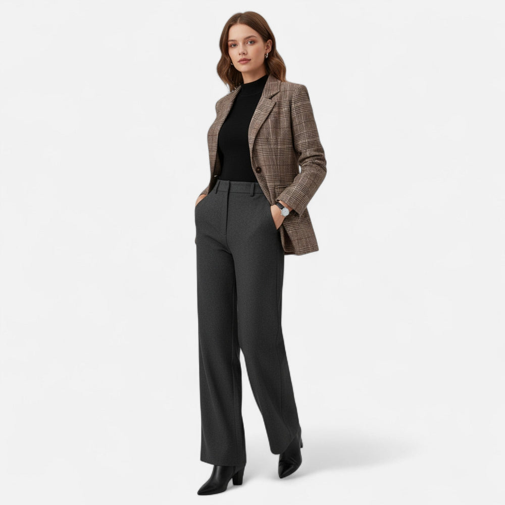 Model toont de geruite blazer in een volledige outfit met zwarte top, antracietgrijze broek met wijde pijpen en zwarte laarzen, handen in de zakken. 2