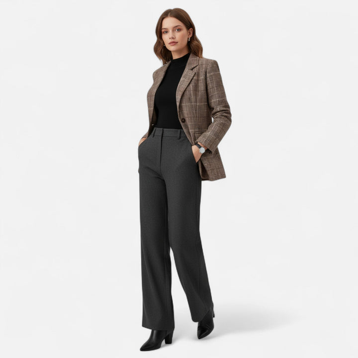 Model toont de geruite blazer in een volledige outfit met zwarte top, antracietgrijze broek met wijde pijpen en zwarte laarzen, handen in de zakken. 2