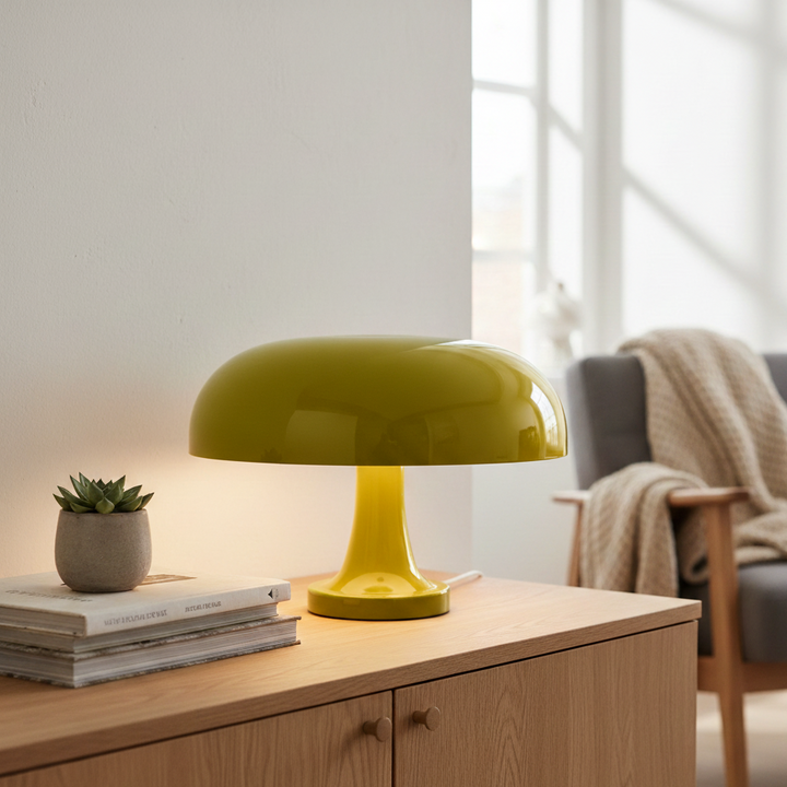 Geelgroene paddenstoellamp met glanzende kap op een houten dressoir, naast een vetplantje en boeken, met een fauteuil op de achtergrond. 11
