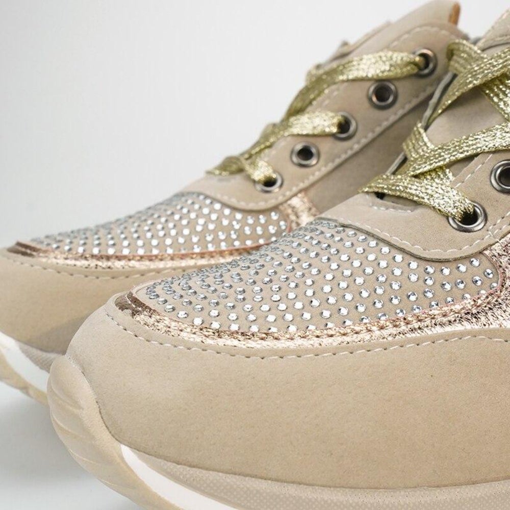 Close-up van de neus van beige sneakers met gouden veters en een vlak vol glinsterende steentjes langs de metallic rand. 10