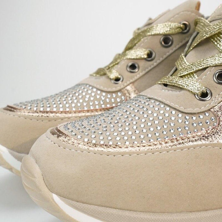 Close-up van de neus van beige sneakers met gouden veters en een vlak vol glinsterende steentjes langs de metallic rand. 10