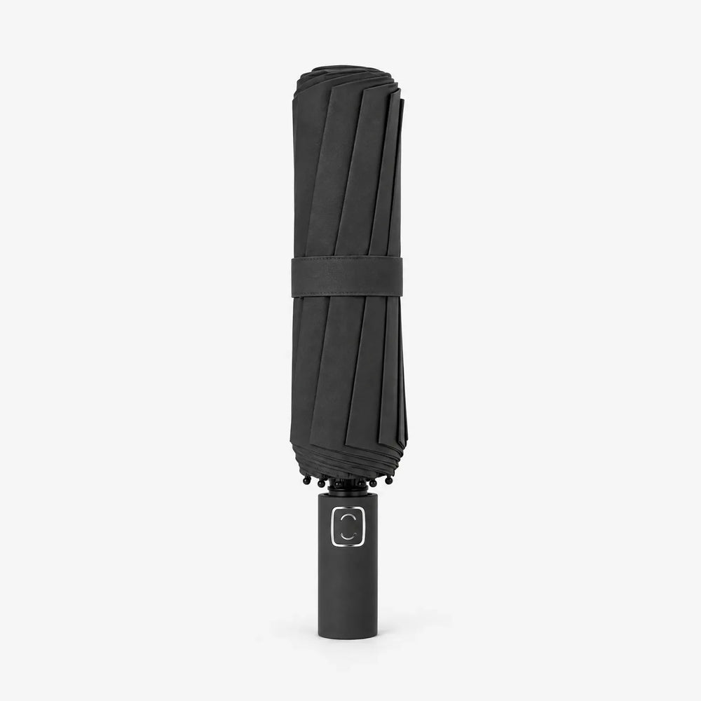 Dichtgevouwen zwarte paraplu met bandje, rechtopstaand op een witte achtergrond, met de handgreep onderaan. 2