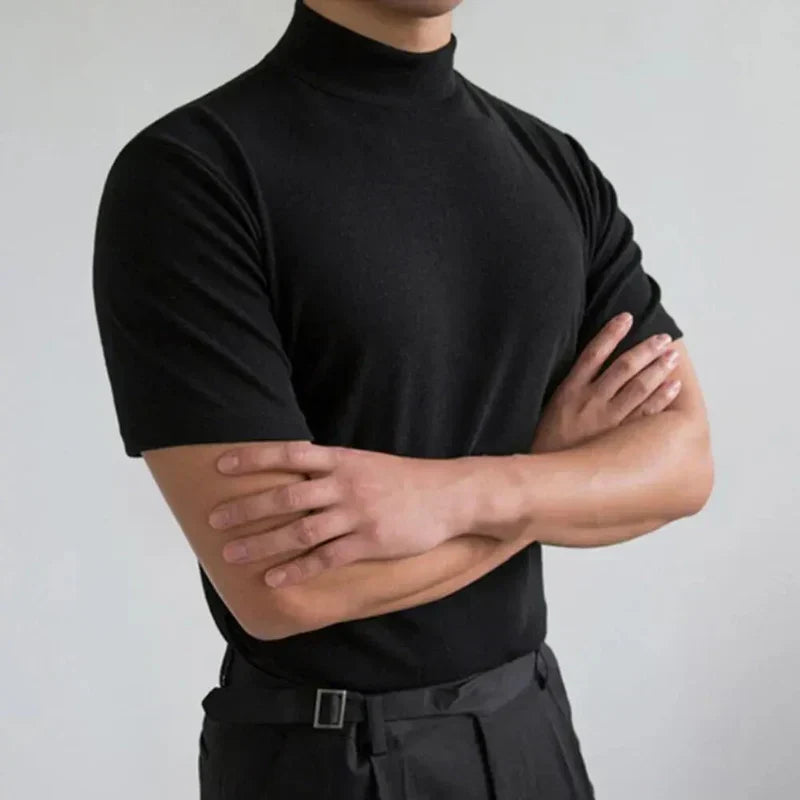 Zwart shirt met opstaande hals en korte mouwen, gedragen door een model met gekruiste armen voor een lichtgrijze muur. 2