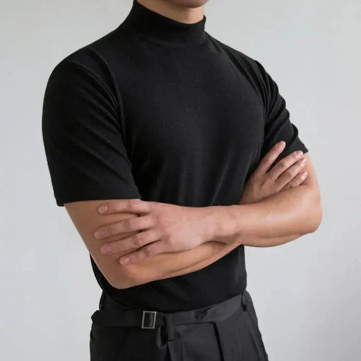 Zwart shirt met opstaande hals en korte mouwen, gedragen door een model met gekruiste armen voor een lichtgrijze muur. 2