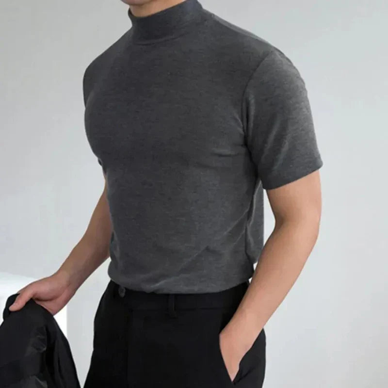 Donkergrijs shirt met opstaande hals en korte mouwen, gedragen door een model met één hand in de zak van een zwarte broek. 3