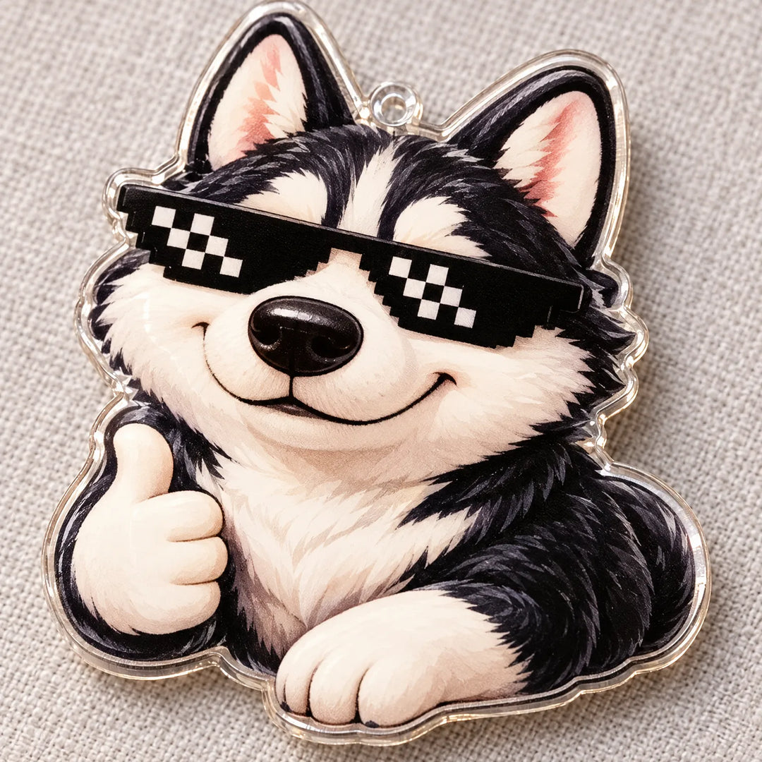 Close-up van een transparante acrylhanger met een lachende husky met pixelzonnebril en een duim omhoog, liggend op een grijze stoffen ondergrond. 4