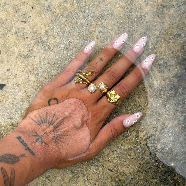 Getatoeëerde hand met bloemetjesnagels in ondiep water, met de gouden hartring aan de ringvinger naast een slangenring en parelringen. 4