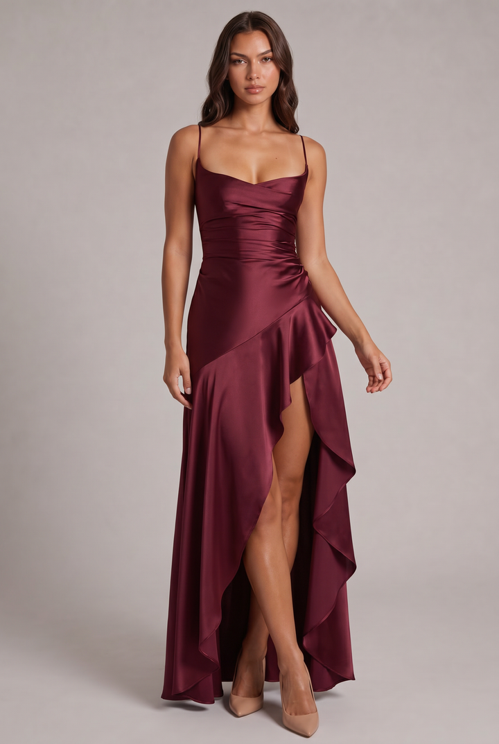Model draagt de satijnachtige maxi jurk in bordeauxrood van voren, met gedrapeerde halslijn, volant en hoge split, gecombineerd met nude pumps. 1