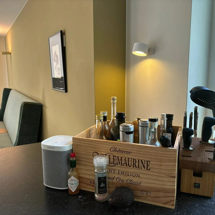 Witte wandlamp brandt in een keukenhoek boven een kist met flessen en kruidenmolens 2
