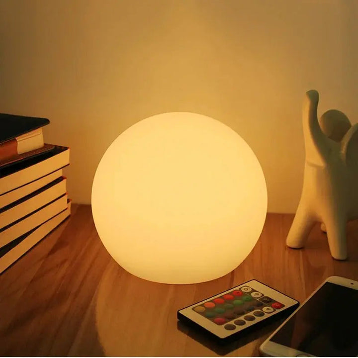 Warm oplichtende ledbol op een houten tafel naast een stapel boeken, met de kleine afstandsbediening en een smartphone ervoor. 1