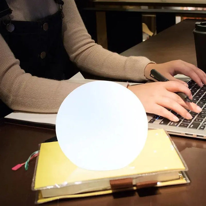 Helder wit oplichtende ledbol op een notitieboek op een bureau, terwijl iemand achter een laptop werkt. 7