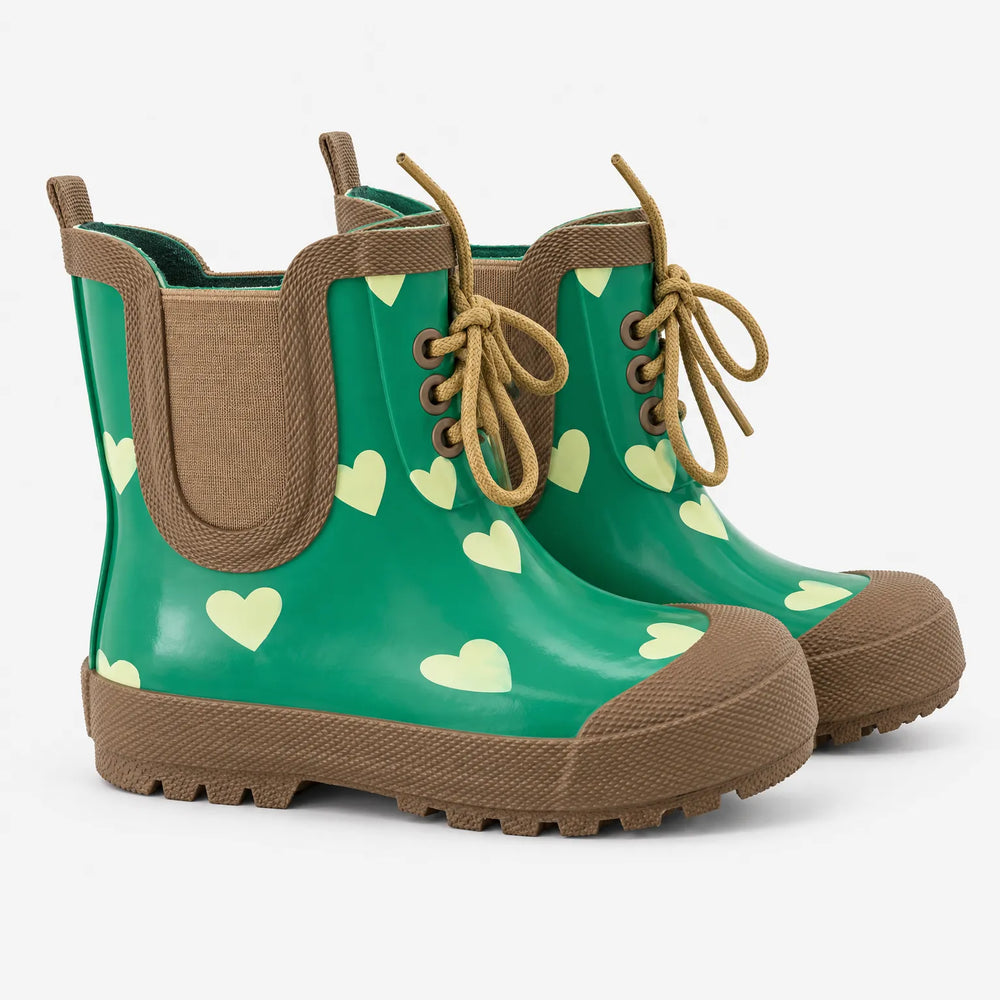 Paar groene rubberen laarzen met lichte hartjes, bruine neus en zool, elastiek aan de zijkant en bruine veters op een witte achtergrond. 2
