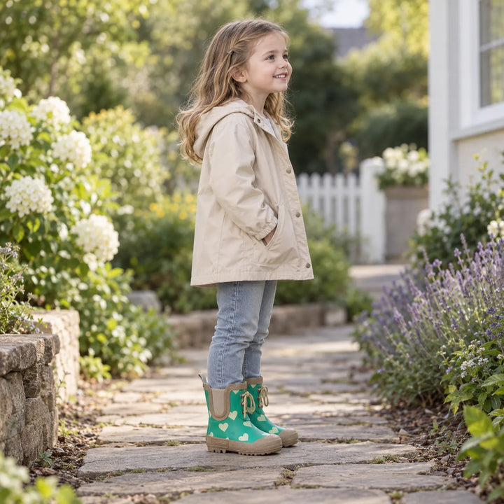 Lachend meisje in beige jas en spijkerbroek draagt de groene hartjeslaarzen op een tuinpad met witte en paarse bloemen. 3