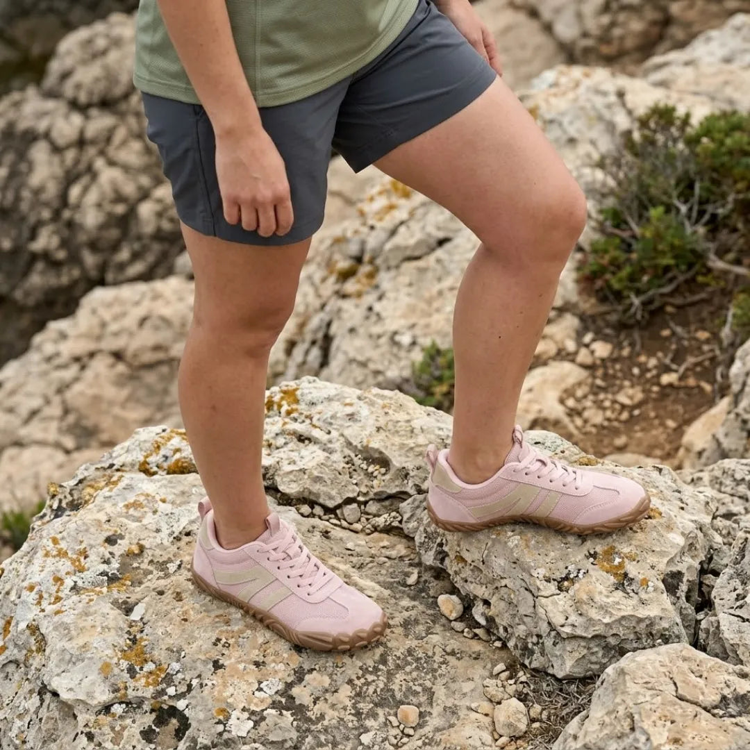 Model in korte broek draagt de roze barefootschoenen en staat met een voet omhoog op een rotsachtige ondergrond. 11