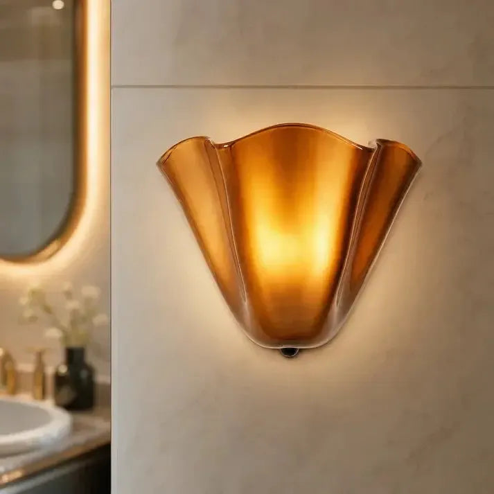 Amberkleurige glazen wandlamp met warm licht tegen een marmerachtige muur, met een badkamerspiegel en wastafel aan de zijkant. 10