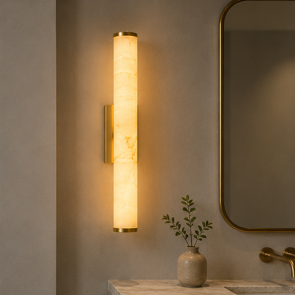 Verticale verlichte wandlamp met marmeren buis en messing uiteinden boven een badkamerwastafel, naast een ronde spiegel en een vaas met takjes. 2