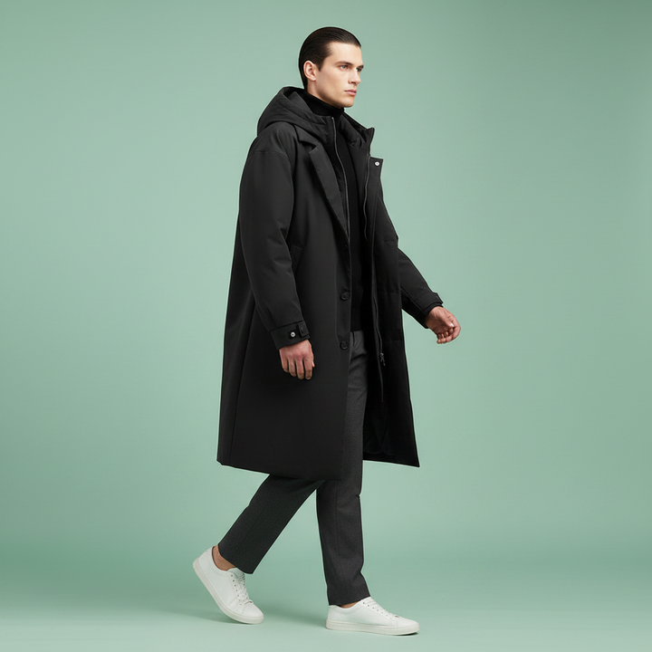 Wandelend model in zwarte lange jas met capuchon, open gedragen met grijze broek en witte sneakers, op groene achtergrond. 7