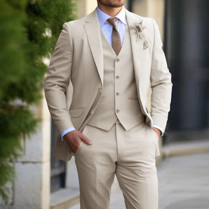 Torso van een man in beige pak met gilet, bruine stropdas en corsage, handen in de zakken 2