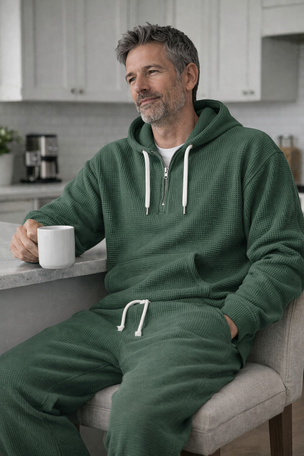 Man in groene set zit aan een keukenblok met een witte mok, hand in de zak van de hoodie. 3