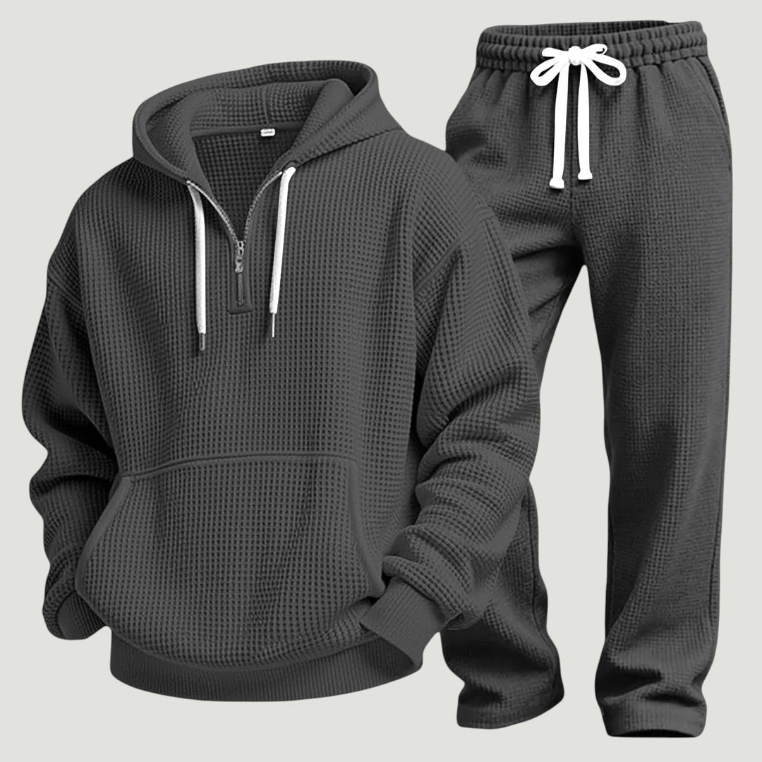 Donkergrijze set uitgelegd: hoodie met halve rits en voorzak naast de joggingbroek met witte koorden. 7