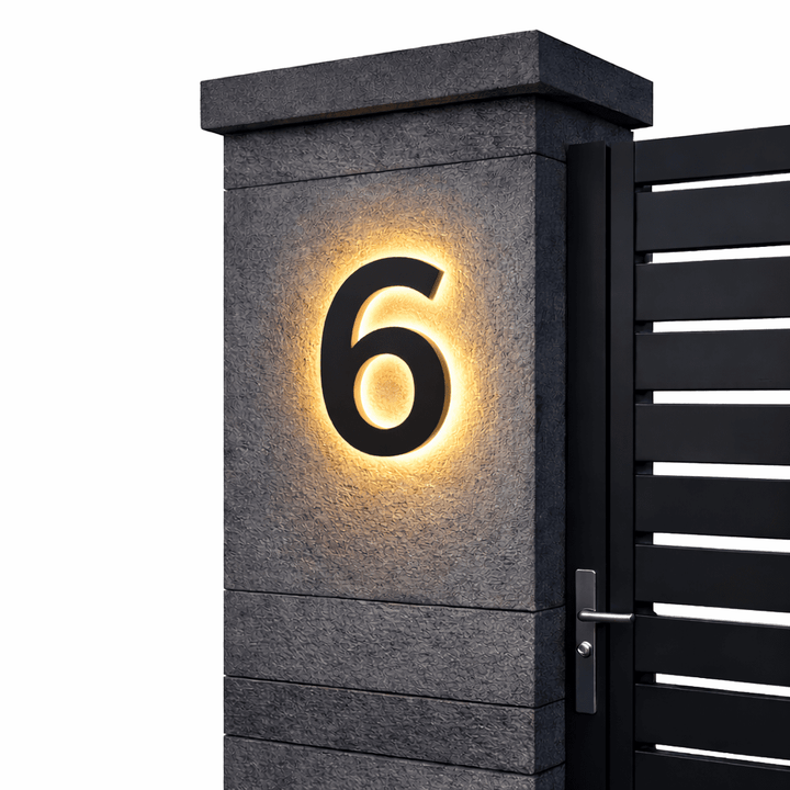Zwart huisnummer 6 met warm achtergrondlicht op een grijze stenen poortpilaar, rechts de zwarte poort met kruk. 12