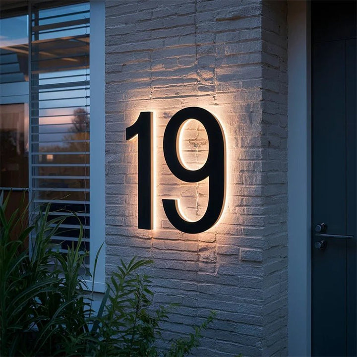 Verlicht zwart huisnummer op een witte bakstenen gevel naast een donkere voordeur, het licht schijnt langs de randen van de cijfers. 4