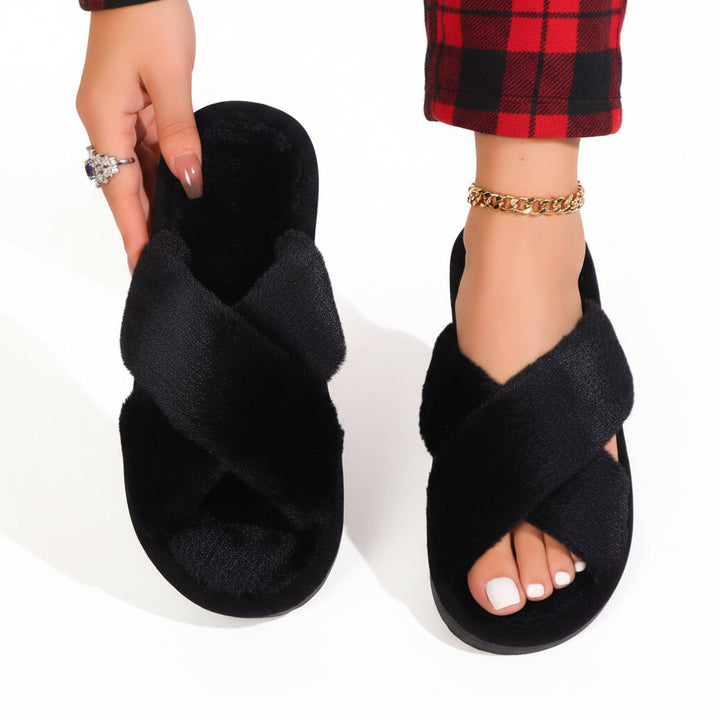 Zwarte pluche slippers met gekruiste banden; het model toont één slipper en draagt de andere bij een rood geruite broek. 9