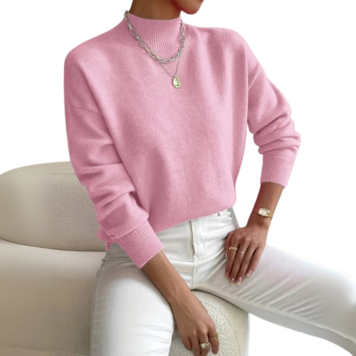 Model zit op een crèmekleurige bouclé stoel in een roze trui met opstaande ribkraag, witte broek en zilveren kettingen. 1